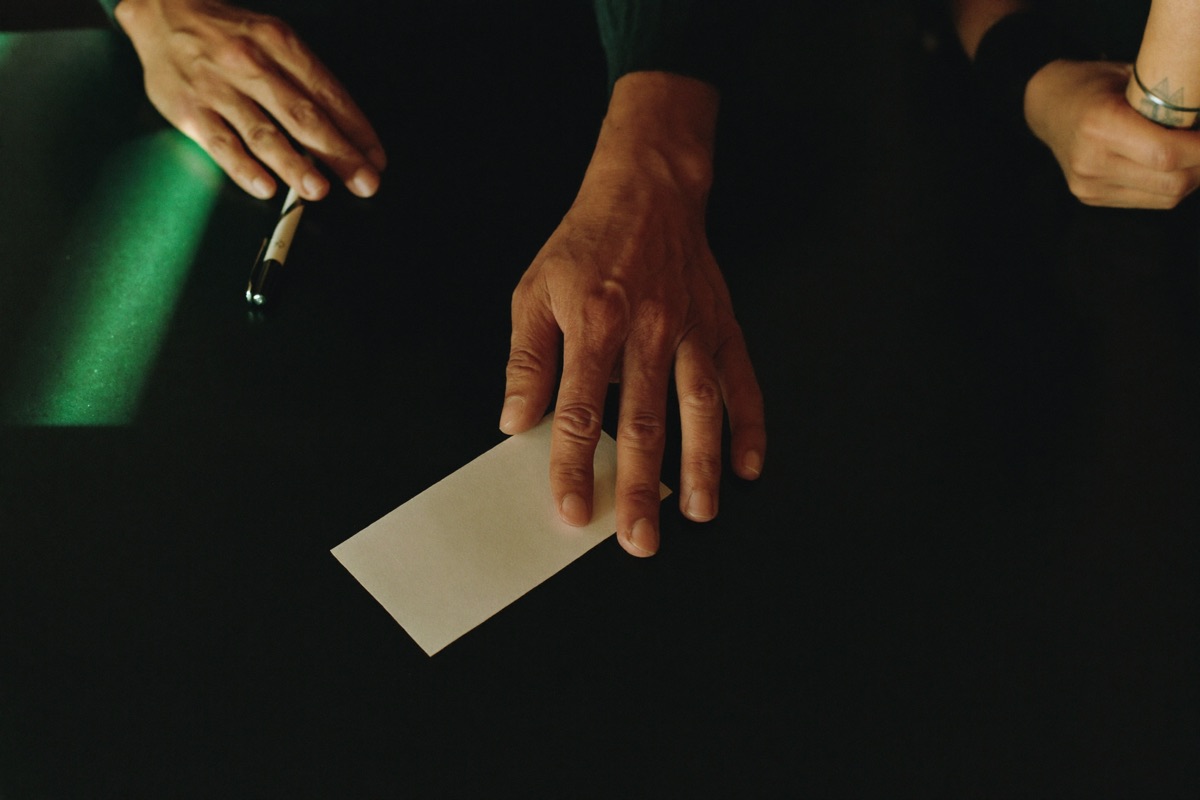 A woman at a numbers counter inside a casino writing four digits on a slip while the clerk keys them in, a draw board showing the next draw date on the wall behind them