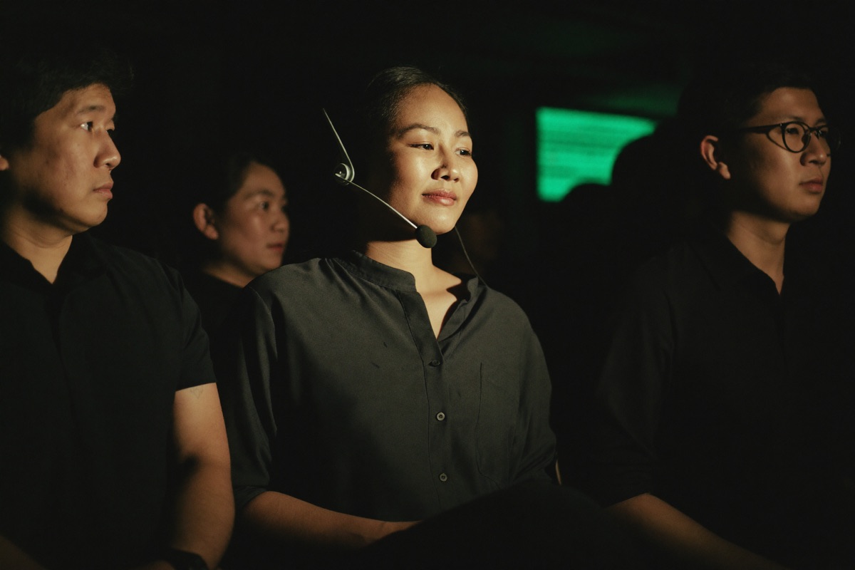 A host at a service desk inside the casino wearing a headset, leaning forward to answer a player standing at the counter