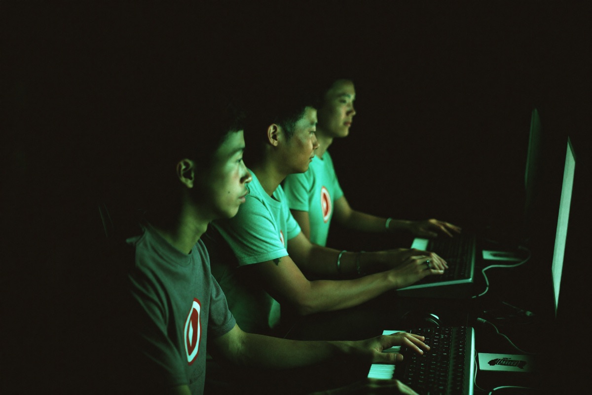Three friends in the e-sports corner of a casino watching a Mobile Legends match on a wide screen, one of them holding a phone with a bet slip open, slot cabinets lit behind them