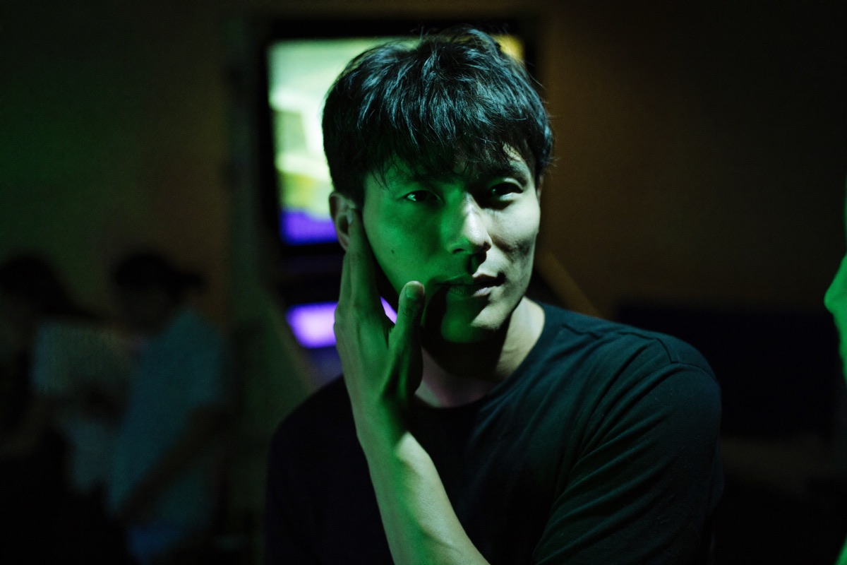 A young man standing at a tall touch terminal in a bright games room inside a casino, one finger on the screen as a rising line chart fills it, two friends watching over his shoulder