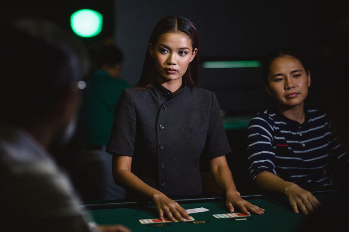 A live baccarat table from the player's side, the dealer in a black waistcoat sliding a card off the shoe while three seated players watch, chips stacked on the felt