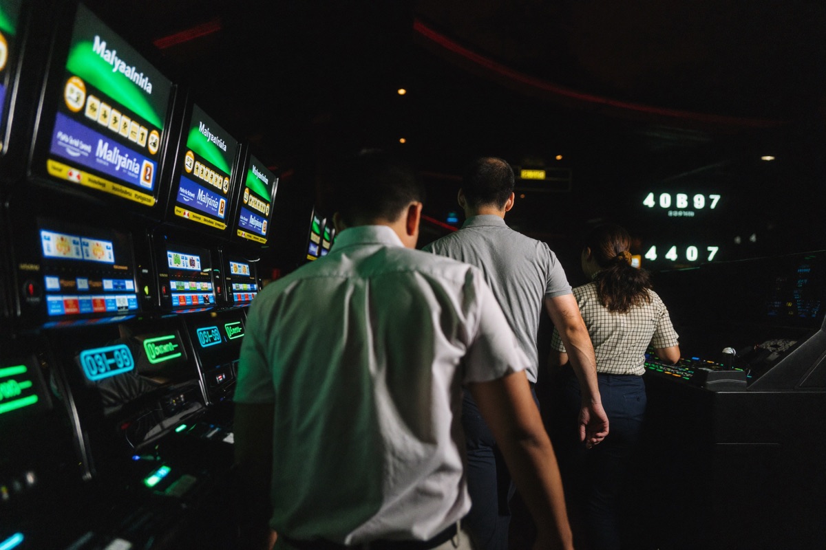 A slot hall at night with three rows of lit cabinets, a woman in a red jacket leaning in to press the spin button on the nearest machine while two men watch her screen