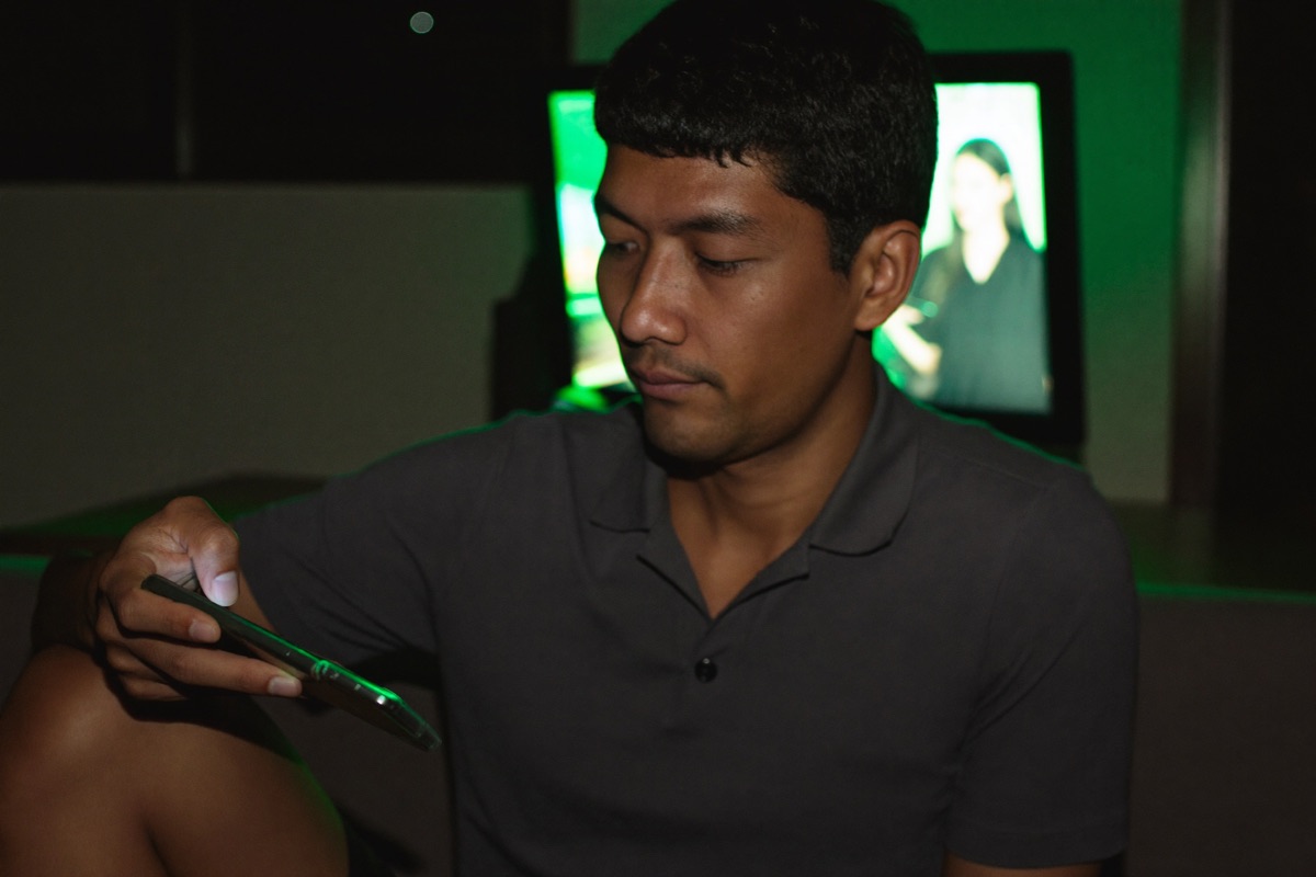 A man in his thirties on a sofa at home holding a laptop with a slot grid on the screen, a phone face up beside him showing the same reels
