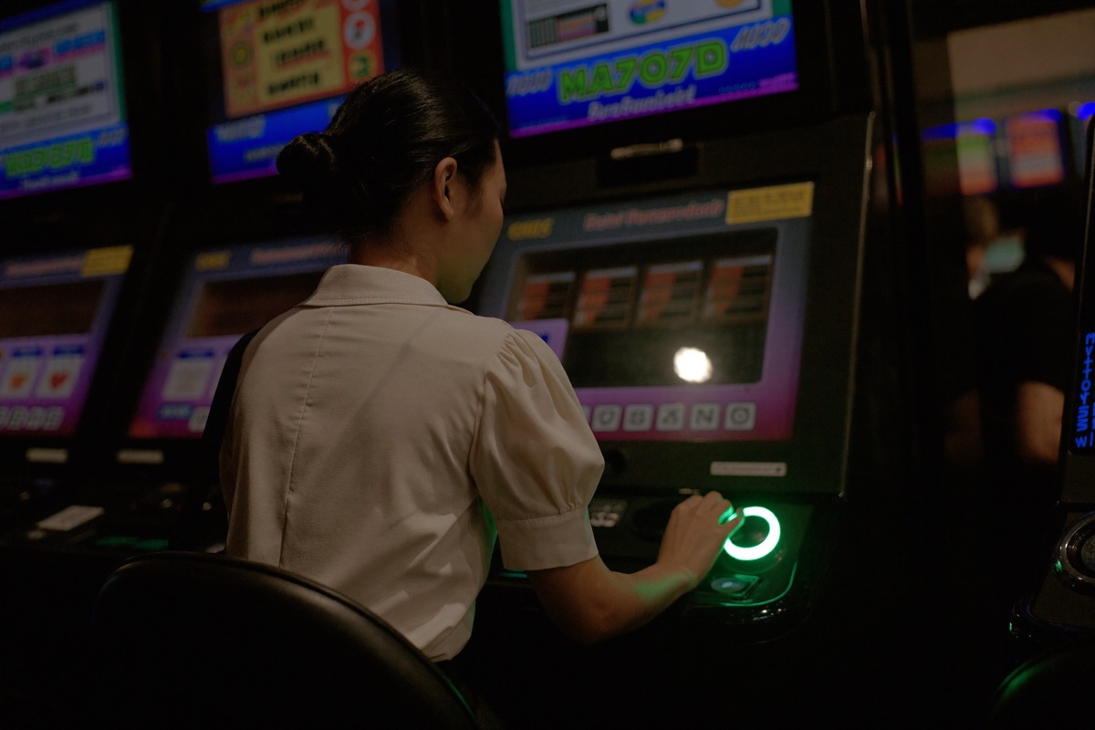 A view over a seated player's shoulder at a tall slot cabinet, five reels mid-spin with a gold symbol lined up, his hand resting on the bet button